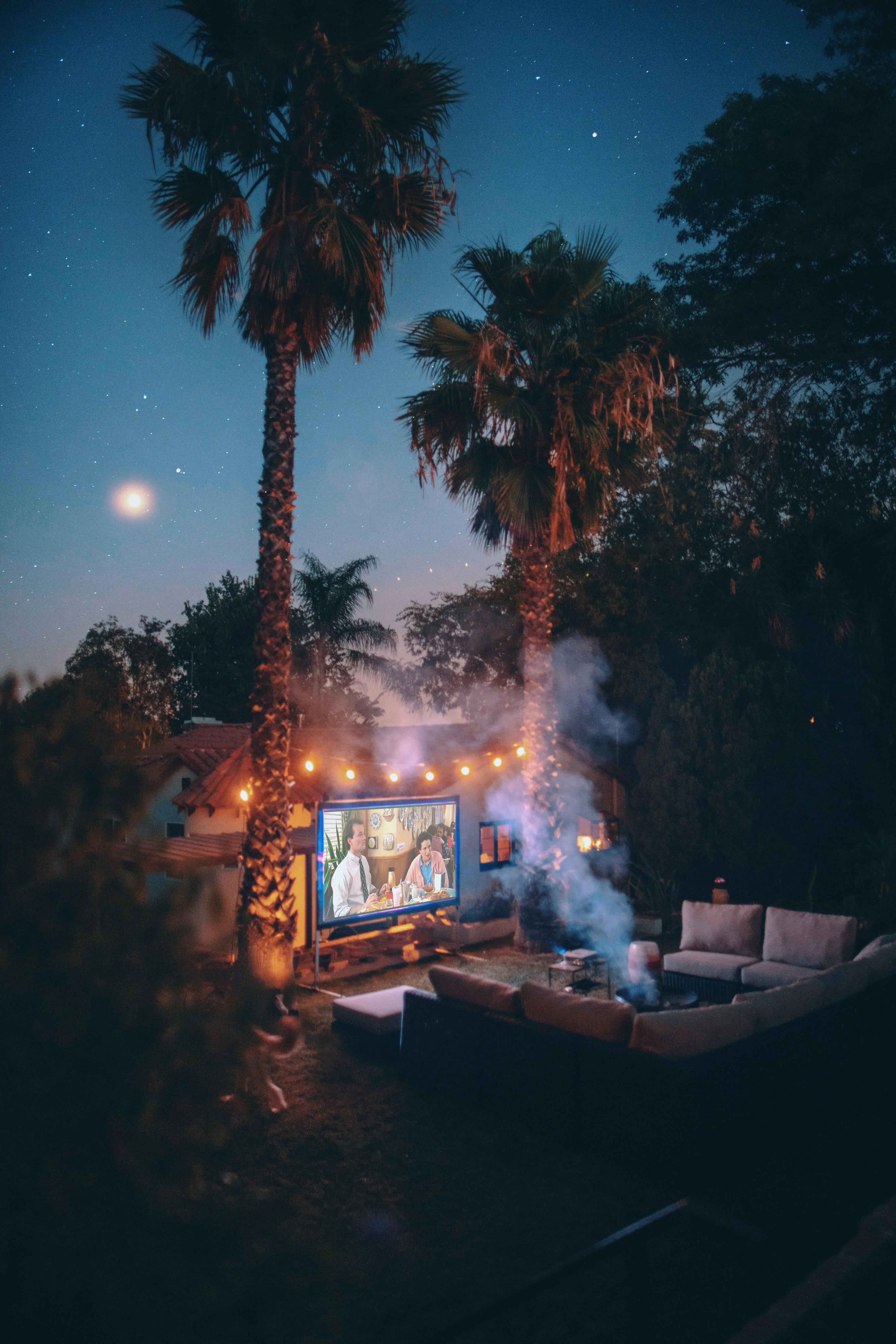 Outdoor evening setting with a movie screen, palm trees, and people watching TV.